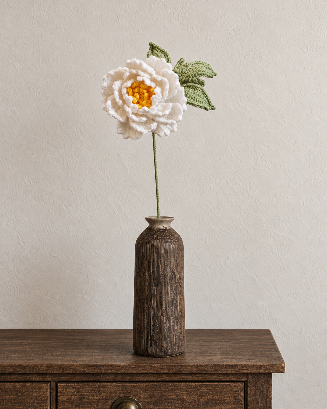 White layered crochet flower with yellow centre and green leaves in a walnut-brown textured vase; vase not included